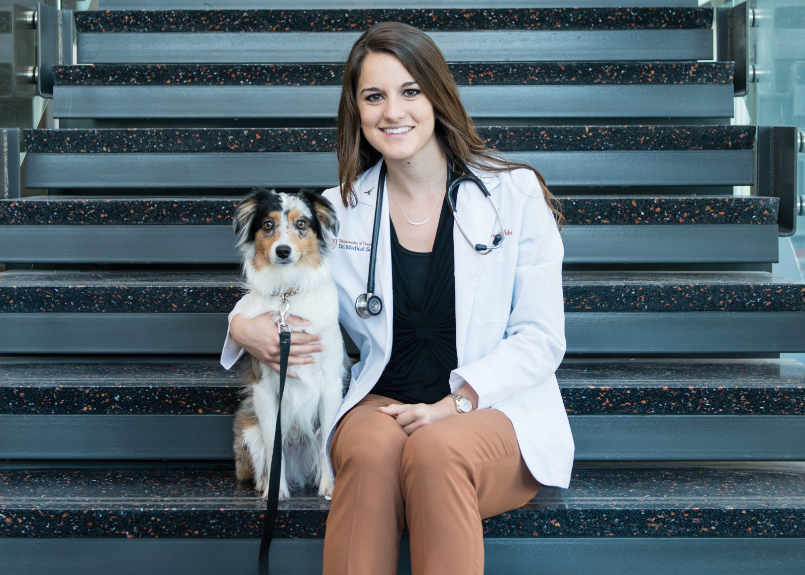 Cierra Grubbs sits on the stairs of Dell Medical School with her arm around her dog. She's wearing a lab coat and stethoscope.