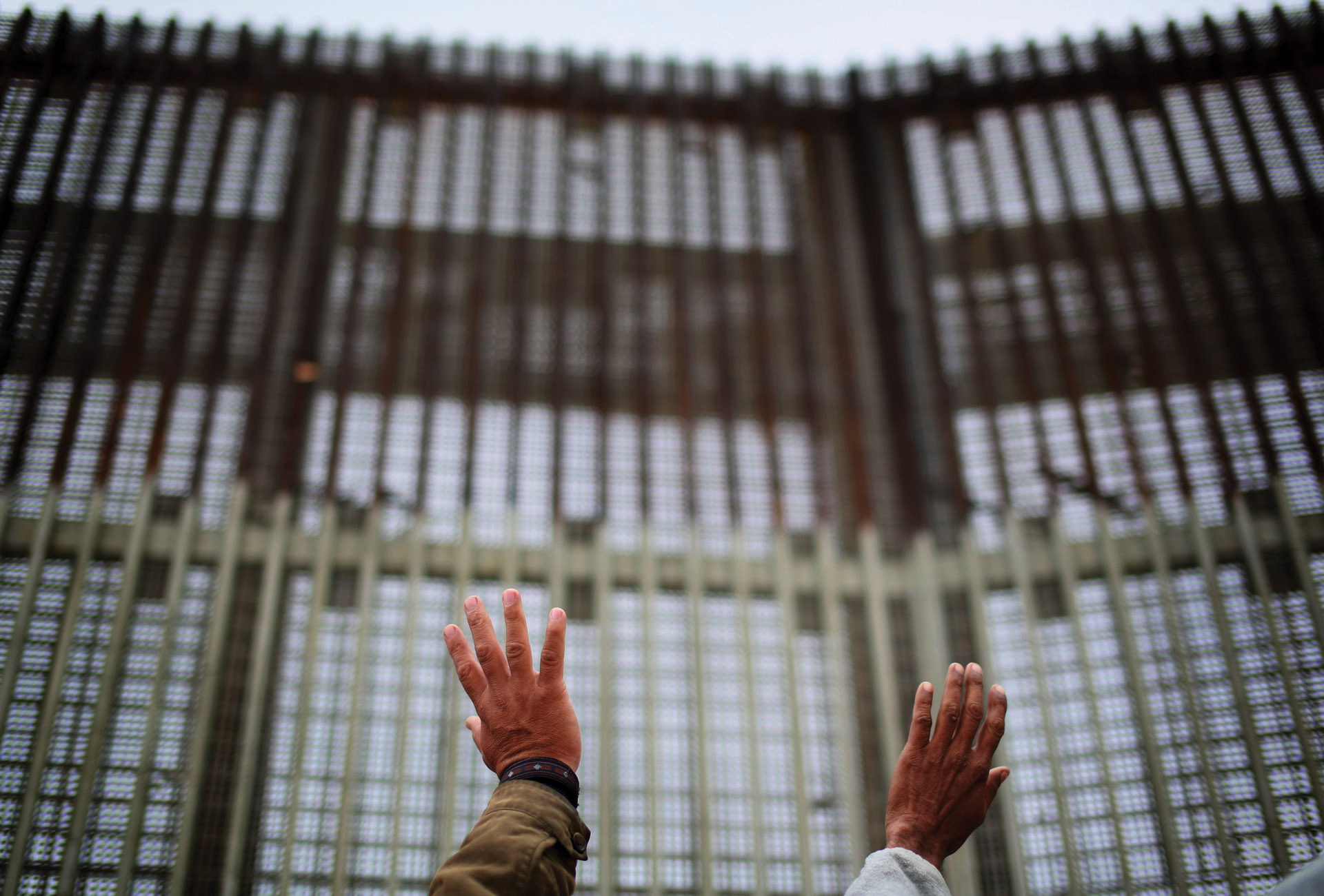 Photo of two outstretched hands in front of a large, imposing border fence.