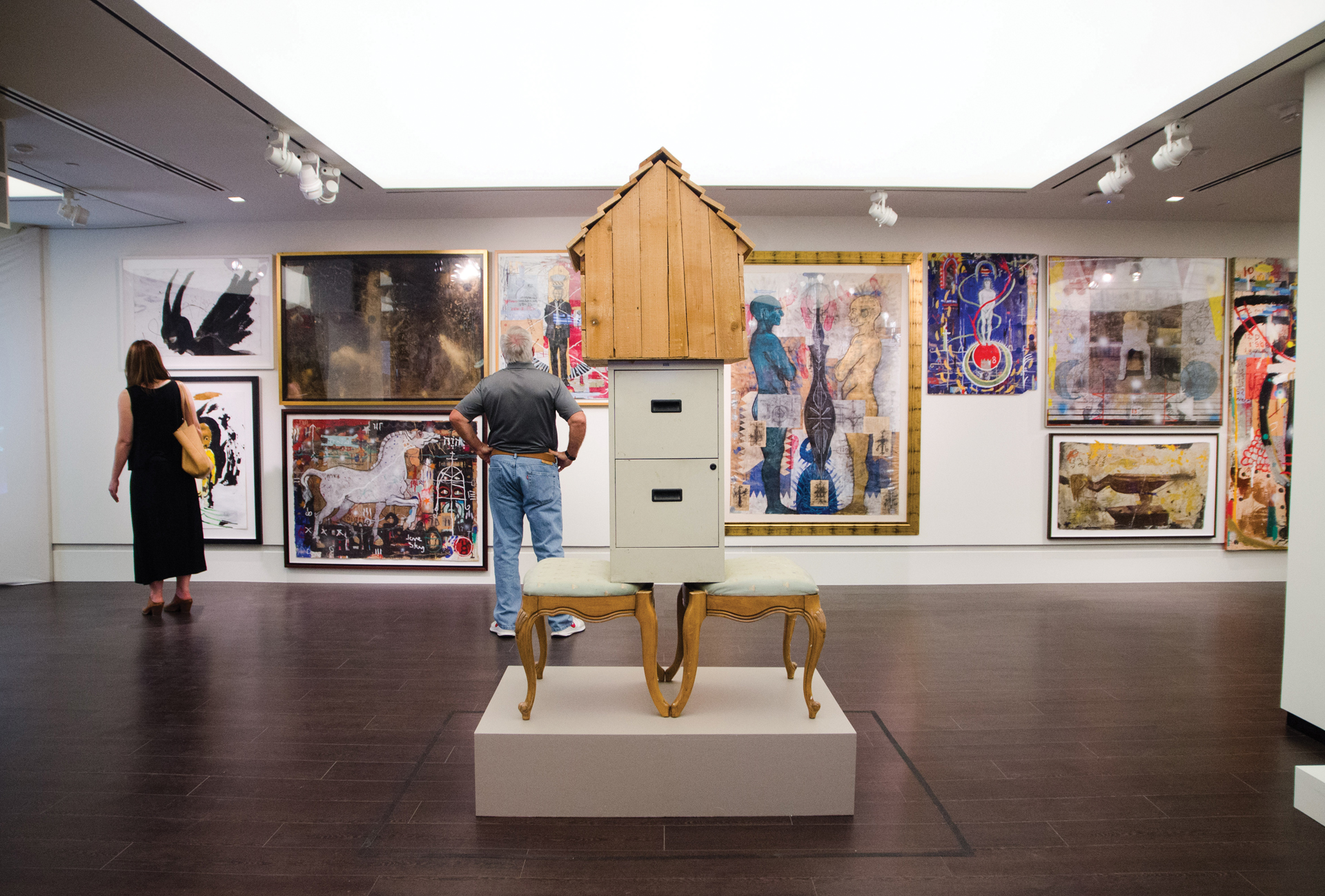 Photo artwork displayed in the Christian-Green Gallery. The piece of artwork in the foreground, titled "The Book I Will Never Write," is two back-facing chairs with a filing cabinet and large birdhouse-like structure on top. Various large-scale paintings can be seen in the background with people looking at them.